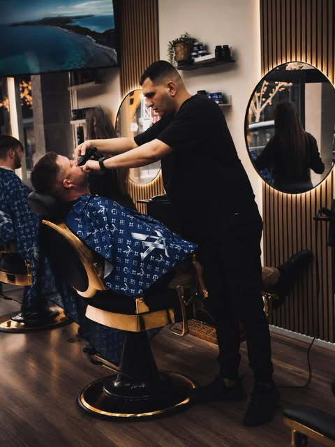 A barber grooming a client in the warm, softly lit LUXURIOUS BARBER ON HAY shop.